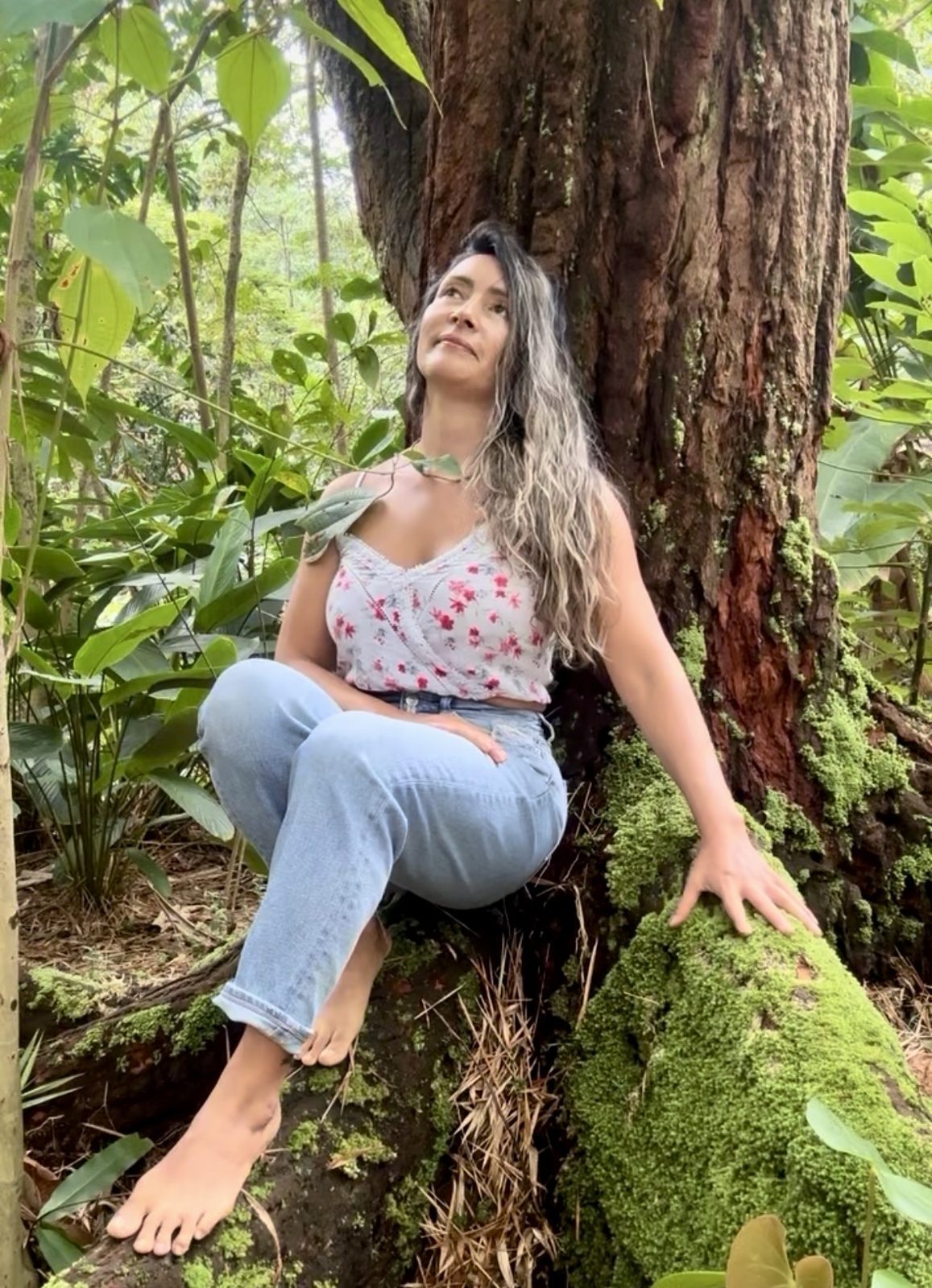 Olga López sentada junto a un árbol en el bosque, mirando hacia arriba con serenidad