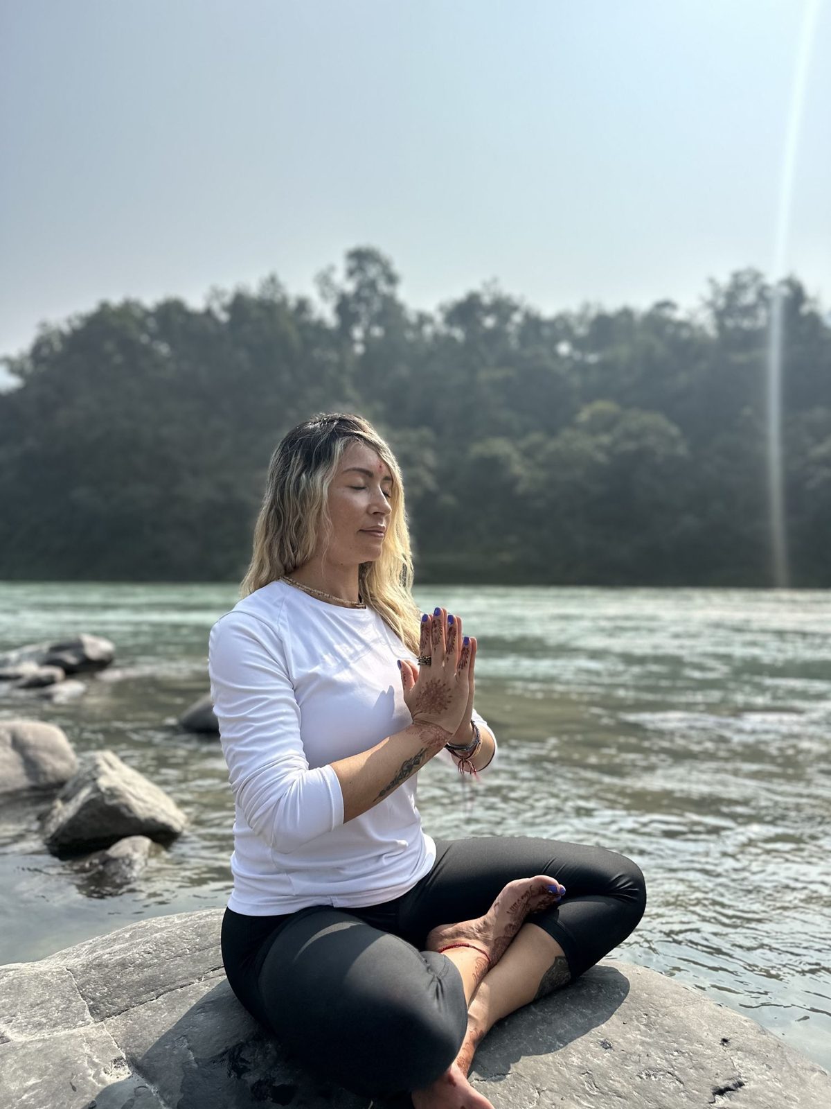 Olga meditando con las manos unidas a la orilla de un río, envuelta en luz